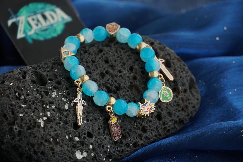 May include: A beaded bracelet featuring faceted light blue stones, gold-toned spacers, and various metal charms including a sword, a rectangular emblem, a colorful mask, and a circular green pendant, with a Zelda logo visible in the background.