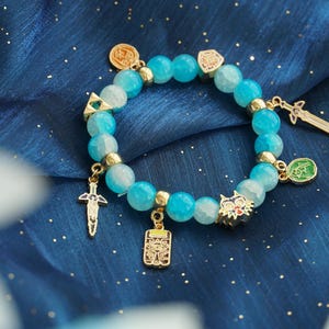 May include: A stretch bracelet featuring round, translucent light blue beads interspersed with small gold-toned metal spacers. The bracelet is adorned with several gold-toned metal charms, including two swords, a triangular emblem, and a wolf head.