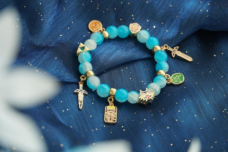 May include: A stretch bracelet featuring round, translucent light blue beads interspersed with small gold-toned metal spacers. The bracelet is adorned with several gold-toned metal charms, including two swords, a triangular emblem, and a wolf head.