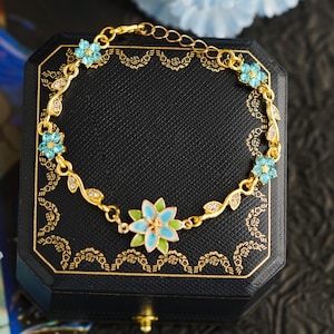 Op de afbeelding: Goudkleurige armband met een bloemmotief, voorzien van lichtblauwe en witte emaille bloemen en heldere kristallen. De armband wordt gepresenteerd op een zwarte sieradendoos met gouden details, die de delicate details benadrukken.