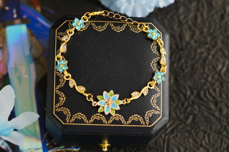 Op de afbeelding: Goudkleurige armband met een bloemmotief, voorzien van lichtblauwe en witte emaille bloemen en heldere kristallen. De armband wordt gepresenteerd op een zwarte sieradendoos met gouden details, die de delicate details benadrukken.