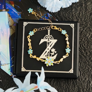 Op de afbeelding: Een goudkleurige armband met een gestileerd zwaard en de letter "Z" met bloemmotieven. De armband is versierd met kleine blauwe bloemvormige edelstenen en wordt gepresenteerd op een zwarte vierkant.