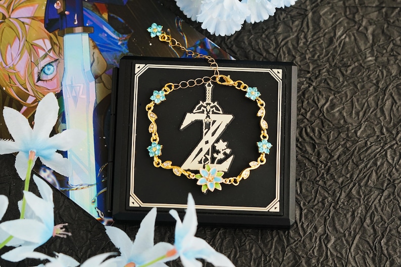 Op de afbeelding: Een goudkleurige armband met een gestileerd zwaard en de letter "Z" met bloemmotieven. De armband is versierd met kleine blauwe bloemvormige edelstenen en wordt gepresenteerd op een zwarte vierkant.