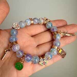May include: A beaded bracelet featuring translucent light blue and purple crackle-effect beads interspersed with gold-toned metal spacers. The bracelet includes several charms, including a shield, a sword, a mask, a green circular emblem, and a triangle.
