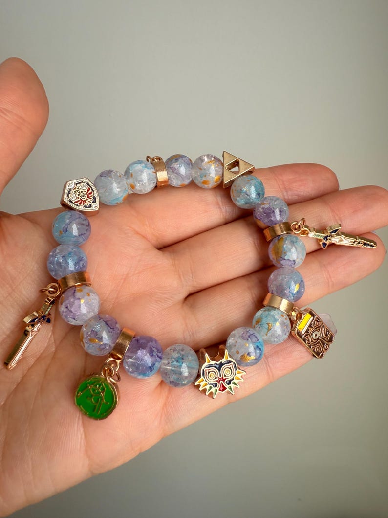 May include: A beaded bracelet featuring translucent light blue and purple crackle-effect beads interspersed with gold-toned metal spacers. The bracelet includes several charms, including a shield, a sword, a mask, a green circular emblem, and a triangle.
