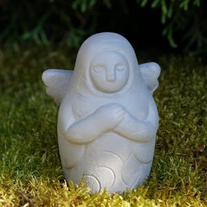Puede incluir: Una figura de ángel de color gris claro con textura suave, cabeza redondeada, ojos cerrados y manos cruzadas sobre el pecho. La pieza presenta pequeñas alas a los lados y un sutil patrón de espirales repetido en la parte inferior del cuerpo.