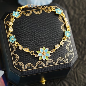 May include: A gold-toned bracelet featuring a floral design with light blue and green enamel accents. The bracelet has small, clear gemstones and is displayed on a black jewelry box with gold trim.