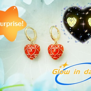 Puede incluir: Pendientes de aro dorados con colgantes rojos en forma de corazón. Los colgantes de corazón tienen un diseño de filigrana decorativo. La imagen también incluye el texto "Surprise!" y "Glow in dark!"