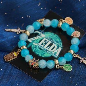 May include: A beaded bracelet featuring light blue faceted beads, gold-toned spacers, and various metal charms including a sword, a mask, and circular emblems. The bracelet is displayed on a black card with The Legend of Zelda Tears of the Kingdom text.