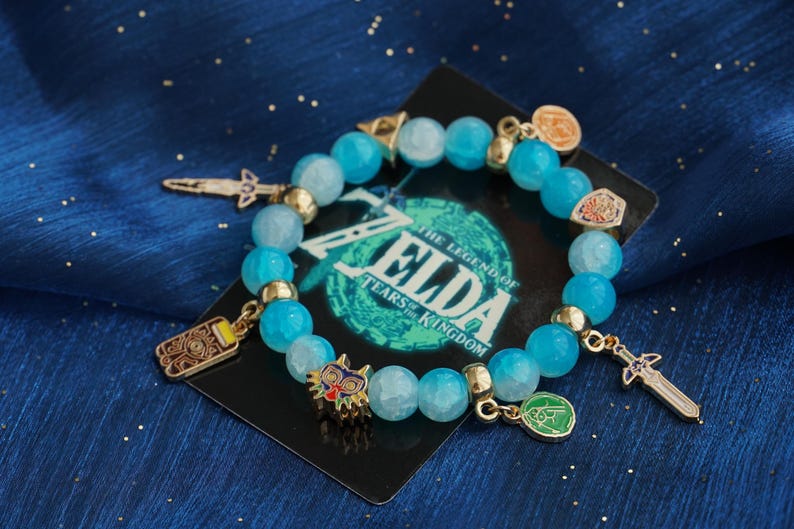May include: A beaded bracelet featuring light blue faceted beads, gold-toned spacers, and various metal charms including a sword, a mask, and circular emblems. The bracelet is displayed on a black card with The Legend of Zelda Tears of the Kingdom text.