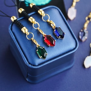 May include: Jewellery charms with gold-coloured accents and colourful gemstone pendants. The charms are attached to black cords. The gemstones are green, red, and blue. The charms are displayed on a blue box against a blue background.