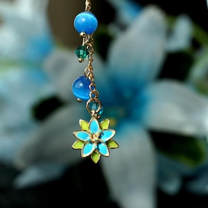 Puede incluir: Una cadena dorada con cuentas azules y verdes y un dije de flor azul. La cadena cuelga de un fondo blanco.