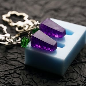 May include: A pair of earrings with purple and green crystal accents. The earrings are on a white silicone mould.