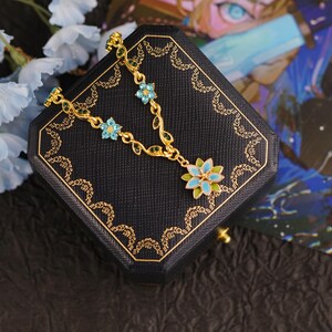 Op de afbeelding: Een goudkleurige ketting met een bloemmotief, met blauwe en groene emaille accenten. De ketting wordt gepresenteerd op een zwarte sieradendoos met gouden randen, naast lichtblauwe bloemen.