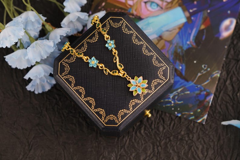 Op de afbeelding: Een goudkleurige ketting met een bloemmotief, met blauwe en groene emaille accenten. De ketting wordt gepresenteerd op een zwarte sieradendoos met gouden randen, naast lichtblauwe bloemen.