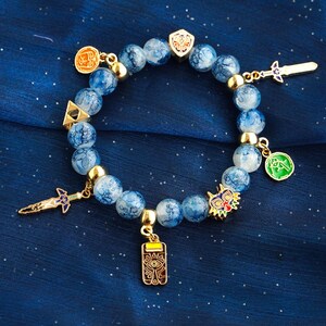 May include: A beaded bracelet featuring round, blue and white marbled beads interspersed with gold-toned metal spacers. The bracelet includes several gold-toned charms, including a sword, a shield, a triangular emblem, a mask, and a rectangular pendant.