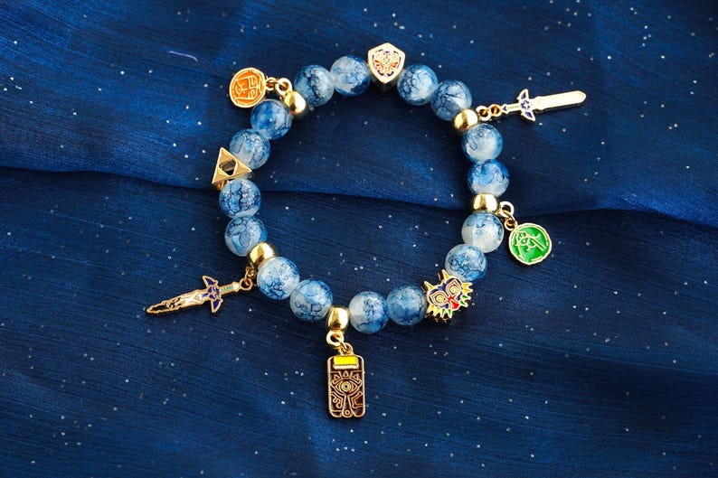 May include: A beaded bracelet featuring round, blue and white marbled beads interspersed with gold-toned metal spacers. The bracelet includes several gold-toned charms, including a sword, a shield, a triangular emblem, a mask, and a rectangular pendant.