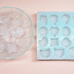 May include: A clear glass bowl filled with ice cubes sits next to a light blue silicone mold with various shield-shaped cavities. The mold is designed for creating decorative ice shapes. The background is a solid light pink.