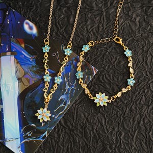 Op de afbeelding: Een goudkleurige sieradenset met een ketting en armband, beide versierd met lichtblauwe bloemen en bladeren. De ketting heeft een hanger met een grotere bloem. De set wordt gepresenteerd op een zwarte, getextureerde ondergrond.