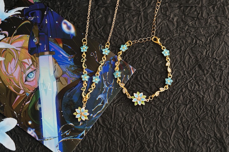 Op de afbeelding: Een goudkleurige sieradenset met een ketting en armband, beide versierd met lichtblauwe bloemen en bladeren. De ketting heeft een hanger met een grotere bloem. De set wordt gepresenteerd op een zwarte, getextureerde ondergrond.