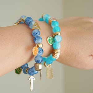 May include: Two beaded stretch bracelets featuring blue crackle glass beads and smooth turquoise-colored beads. The bracelets are accented with gold-toned metal spacers and various small charms, including a sword, a shield, and circular emblems.