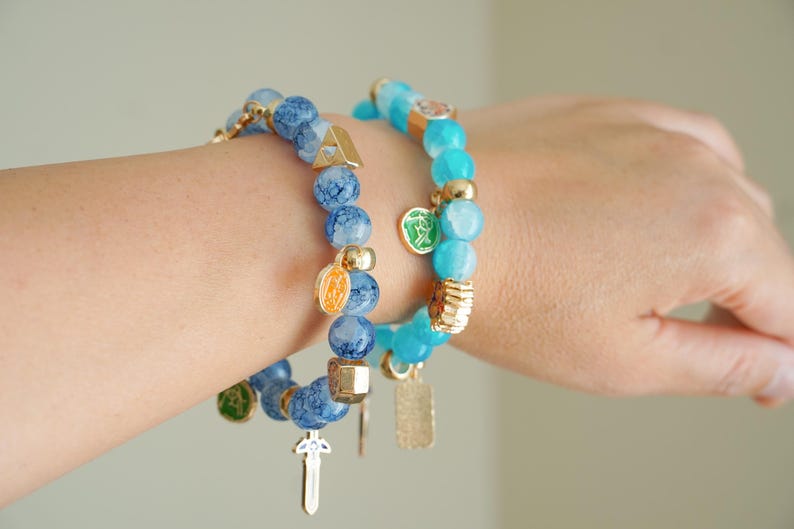 May include: Two beaded stretch bracelets featuring blue crackle glass beads and smooth turquoise-colored beads. The bracelets are accented with gold-toned metal spacers and various small charms, including a sword, a shield, and circular emblems.