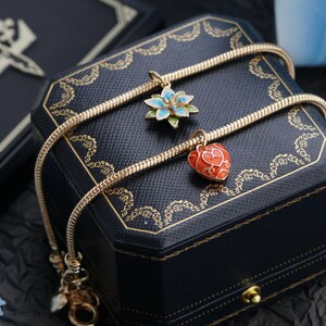 以下が含まれることがあります： 金色のネックレス。2つのチャームがあり、1つは青と緑の花、もう1つは赤いハート。ネックレスは、金のトリムが付いた黒いジュエリーボックスの上にかけられています。背景は暗いです。