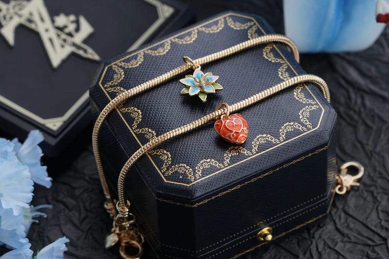 以下が含まれることがあります： 金色のネックレス。2つのチャームがあり、1つは青と緑の花、もう1つは赤いハート。ネックレスは、金のトリムが付いた黒いジュエリーボックスの上にかけられています。背景は暗いです。