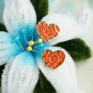 Könnte beinhalten: Nahaufnahme von zwei herzförmigen Ohrringen mit orangefarbenem Filigranmuster, gefasst in goldfarbenem Metall. Die Ohrringe liegen auf einer weiß-blauen Blume mit grünen Akzenten, was ein romantisches und dekoratives Thema ergibt.