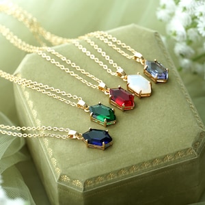 May include: Five gold necklaces with different coloured gemstone pendants. The pendants are all in the shape of a hexagon and are arranged in a row on a green velvet box. The gemstones are blue, white, red, green, and dark blue.