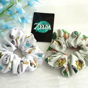 May include: Two white scrunchies with colourful floral and cartoon designs. One features blue flowers and other designs, the other has a light grey background with cartoon characters. A black card with "ZELDA TEARS OF THE KINGDOM" is between them.