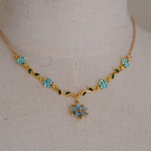 Op de afbeelding: Goudkleurige ketting met een bloemenontwerp. De ketting heeft lichtblauwe, bloemvormige edelstenen en groene bladeren. Een grotere bloemhanger hangt in het midden. De ketting wordt getoond op een mannequin.