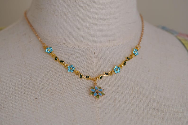 Op de afbeelding: Goudkleurige ketting met een bloemenontwerp. De ketting heeft lichtblauwe, bloemvormige edelstenen en groene bladeren. Een grotere bloemhanger hangt in het midden. De ketting wordt getoond op een mannequin.