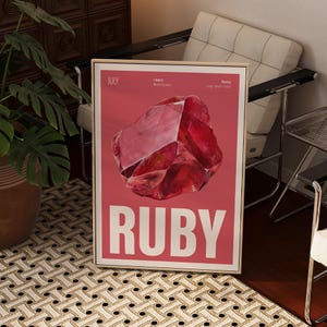Może przedstawiać: Oprawiony wydruk artystyczny z dużą, abstrakcyjną ilustracją rubinu na różowym tle. Słowo "RUBY" jest wyświetlane pogrubioną, białą czcionką na dole. Wydruk jest w złotej ramie.
