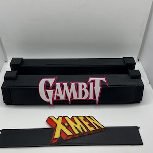 May include: Black 3D-printed display stand with the word "GAMBIT" in white and pink lettering. Below is a black stand with the "X-MEN" logo in yellow, red, and orange. The items are likely for a comic book or superhero fan.