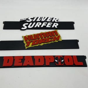 May include: Three black rectangular nameplates with raised lettering. The top plate reads "THE SILVER SURFER" in white. The middle plate reads "HUMAN TORCH" in red and yellow. The bottom plate reads "DEADPOOL" in red with a Deadpool logo.