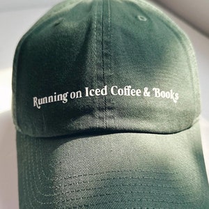 May include: A dark green baseball cap with the text "Running on Iced Coffee & Books" in white. The cap has a curved brim and a fabric strap closure. The hat is made of a soft, durable material.