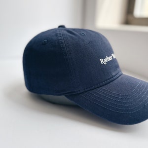 May include: A navy blue baseball cap with a curved peak. The cap has the word "Rather" embroidered in white on the front. The hat is made of a sturdy fabric and has a classic, casual style.
