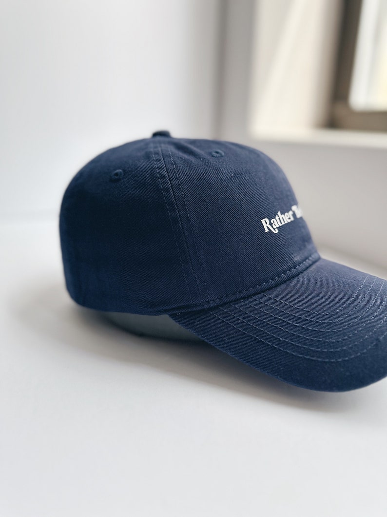 May include: A navy blue baseball cap with a curved peak. The cap has the word "Rather" embroidered in white on the front. The hat is made of a sturdy fabric and has a classic, casual style.