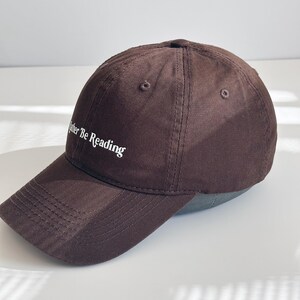 May include: A brown baseball cap with the text "Rather Be Reading" embroidered in white. The cap features a curved peak and a classic six-panel design. The cap is made of a soft, durable fabric.
