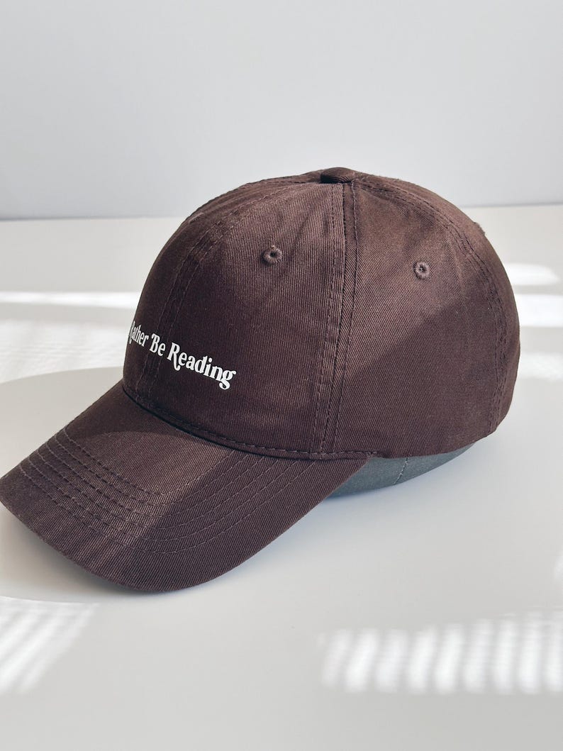 May include: A brown baseball cap with the text "Rather Be Reading" embroidered in white. The cap features a curved peak and a classic six-panel design. The cap is made of a soft, durable fabric.