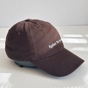 May include: A brown baseball cap featuring the text "Rather Be Reading" in white. The cap has a curved peak and a rounded crown. The cap is made of a soft, textured material. A casual accessory.