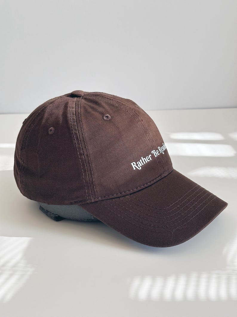 May include: A brown baseball cap featuring the text "Rather Be Reading" in white. The cap has a curved peak and a rounded crown. The cap is made of a soft, textured material. A casual accessory.