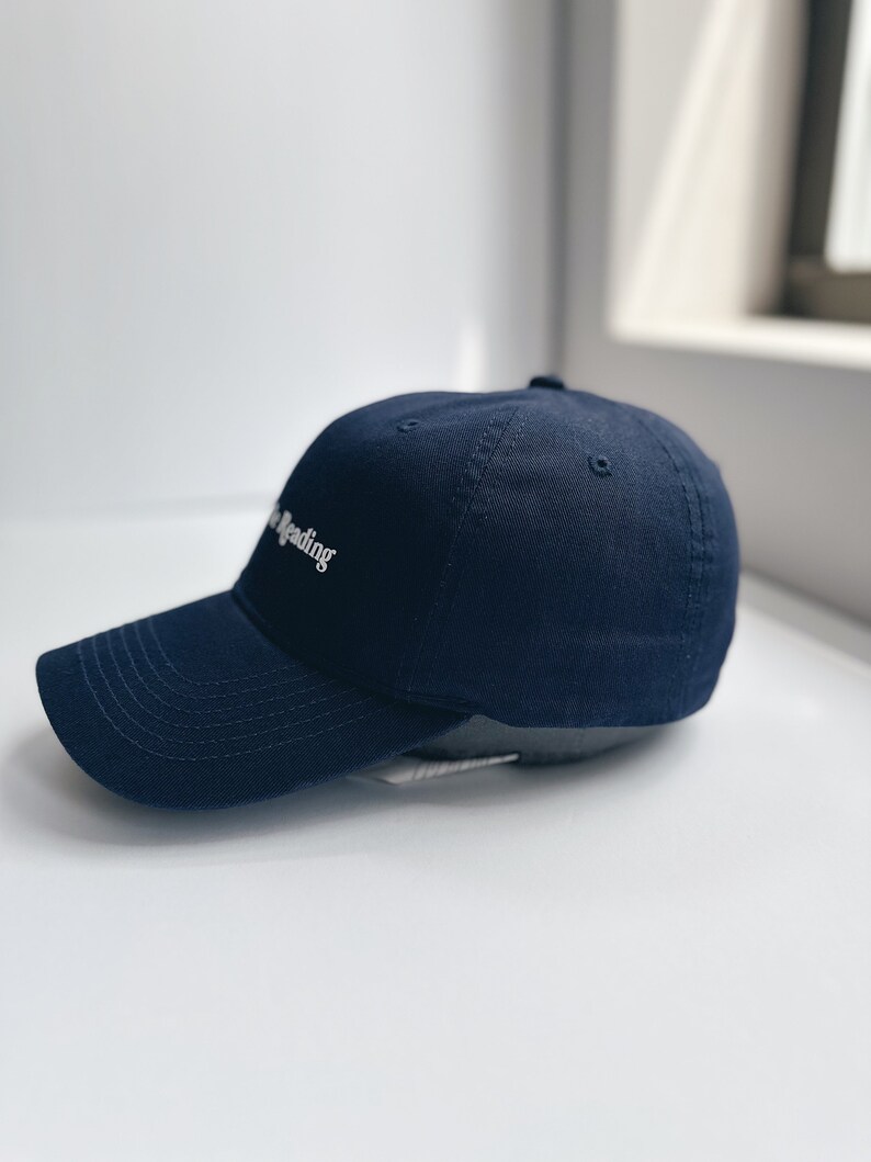 May include: A navy blue baseball cap with a curved peak. The cap has the white text "the reading" embroidered on the front. The hat is made of a soft fabric and has a classic, casual style.