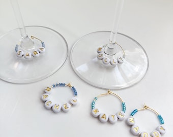 May include: Wine glass charms with names spelled out in gold letters on white beads. The charms are attached to gold hoops and accented with blue beads. The names include Sara, Molly, Rachel, Katie, and Daddy.