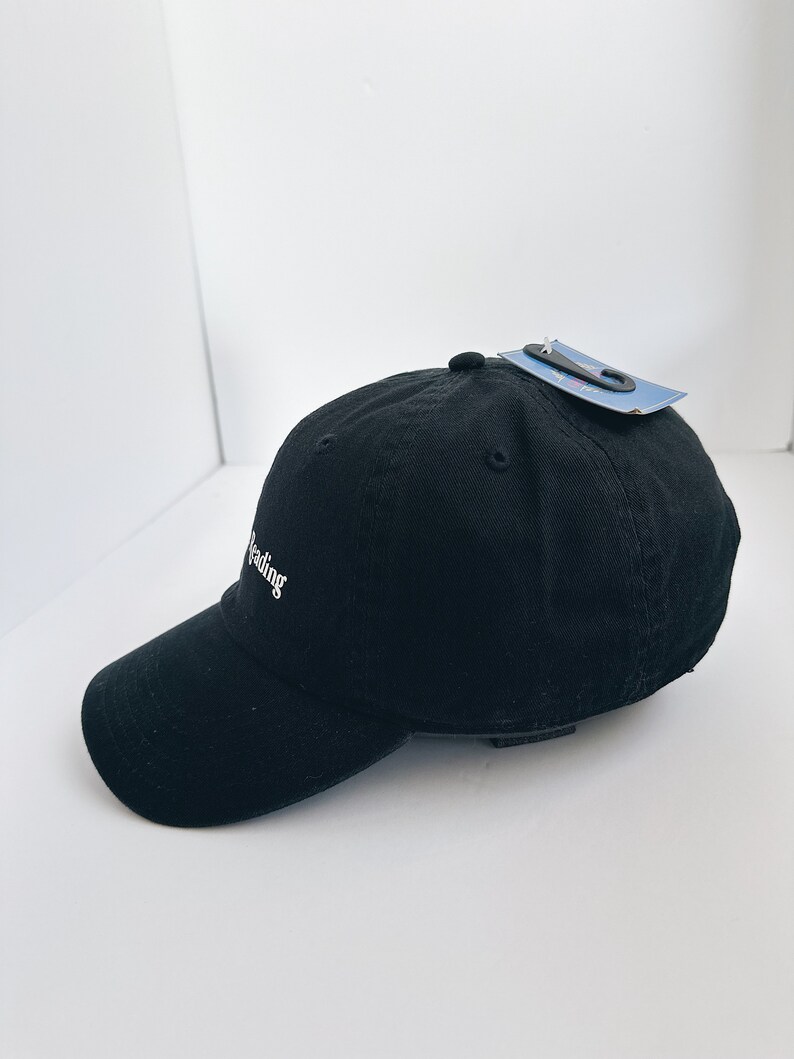 May include: Black baseball cap with a curved peak. The cap features the word "reading" embroidered in white script. A tag is attached to the top of the cap.