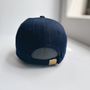 May include: A navy blue baseball cap with a curved peak. The back of the cap has an adjustable strap with a gold-coloured metal clasp. The cap is made of a smooth fabric and has a small button on top.