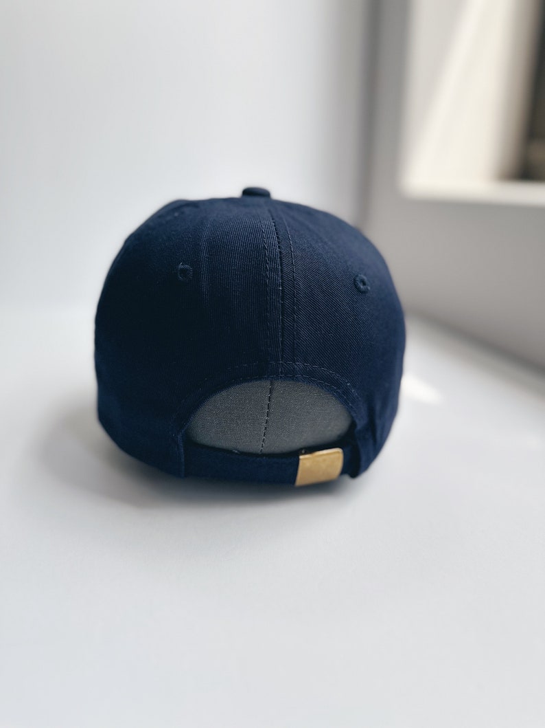 May include: A navy blue baseball cap with a curved peak. The back of the cap has an adjustable strap with a gold-coloured metal clasp. The cap is made of a smooth fabric and has a small button on top.