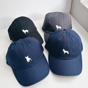 May include: Four baseball caps in black, gray, and navy blue. Each cap features a white dog silhouette on the front. The caps are made of a soft fabric and have a curved brim. The caps are arranged on a white surface.