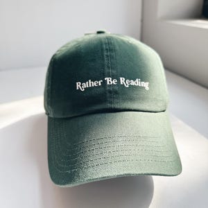 May include: A dark green baseball cap with the text "Rather Be Reading" in white script. The cap is made of a soft fabric and has a curved brim. The hat is a classic style.